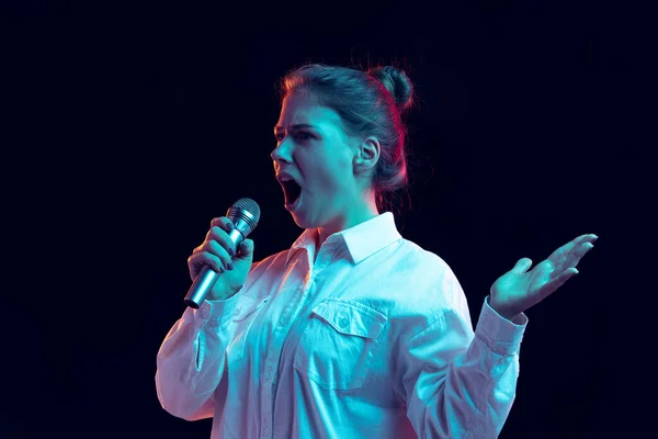 Halve lengte portret van jong mooi meisje in wit shirt met microfoon geïsoleerd op donkere achtergrond in blauw neon licht, filter. Concept van emoties, mode, jeugd — Stockfoto