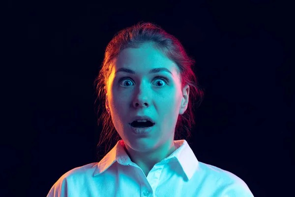 Geschokt jong meisje in wit shirt kijkend naar camera met open mond geïsoleerd op donkere achtergrond in blauw neon licht, filter. Concept van emoties, mode, jeugd — Stockfoto