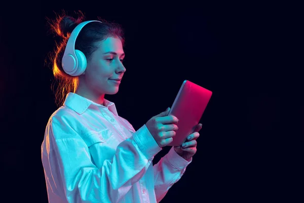 Portrait of young beautiful happy girl in headphones using tablet isolated on orange background in neon light, filter. Concept of emotions, music — Stock Photo, Image