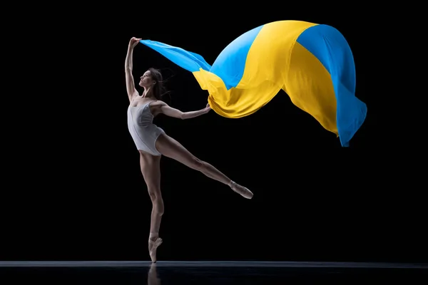 Молода витончена класична балерина танцює з тканиною, пофарбованою синіми і жовтими кольорами прапора України на темному студійному тлі. Мистецтво, мир, свобода і права — стокове фото