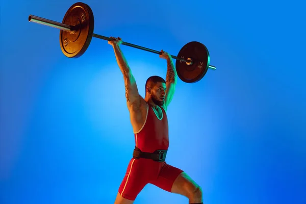 Studio shot di giovane uomo in abbigliamento sportivo rosso che si esercita con bilanciere isolato sfondo blu al neon. Sport, sollevamento pesi, potenza, concetto di risultati — Foto Stock