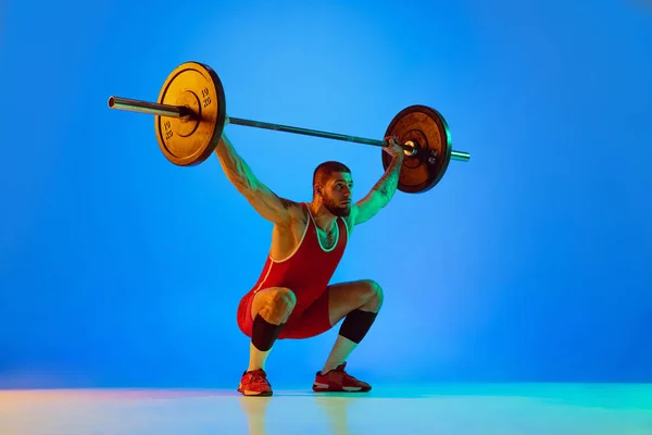 Студійний знімок молодого чоловіка в червоному спортивному одязі, вправи з барбелом ізольовані на синьому фоні в неоні. Спорт, важка атлетика, сила, концепція досягнень — стокове фото