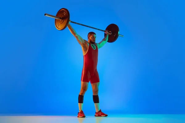 Studio shot van jonge man in rode sportkleding oefenen met lange halter geïsoleerde blauwe achtergrond in neon. Sport, gewichtheffen, kracht, prestaties concept — Stockfoto