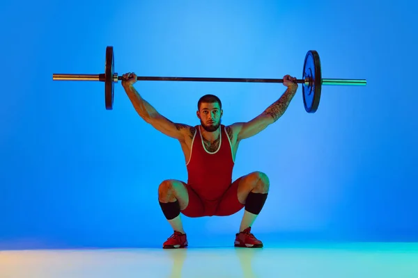 Studio shot van jonge man in rode sportkleding oefenen met lange halter geïsoleerde blauwe achtergrond in neon. Sport, gewichtheffen, kracht, prestaties concept — Stockfoto