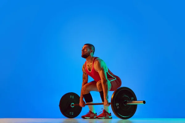 Studio shot di giovane uomo in abbigliamento sportivo rosso che si esercita con bilanciere isolato sfondo blu al neon. Sport, sollevamento pesi, potenza, concetto di risultati — Foto Stock