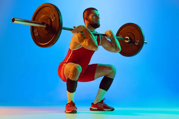 Estudio de tiro de un joven en ropa deportiva de color rojo haciendo ejercicio con la barra de fondo azul aislado en neón. Deporte, levantamiento de pesas, poder, concepto de logros —  Fotos de Stock