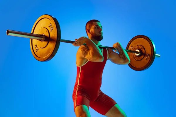 Studio shot van jonge man in rode sportkleding oefenen met lange halter geïsoleerde blauwe achtergrond in neon. Sport, gewichtheffen, kracht, prestaties concept — Stockfoto