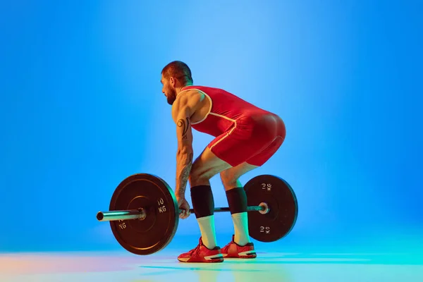 Studyjne ujęcie młodego człowieka w czerwonej odzieży sportowej ćwiczącego ze sztangą odizolowanym niebieskim tle w neonie. Sport, podnoszenie ciężarów, siła, koncepcja osiągnięć — Zdjęcie stockowe