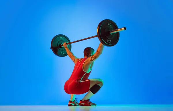 Estudio de tiro de un joven en ropa deportiva de color rojo haciendo ejercicio con la barra de fondo azul aislado en neón. Deporte, levantamiento de pesas, poder, concepto de logros —  Fotos de Stock