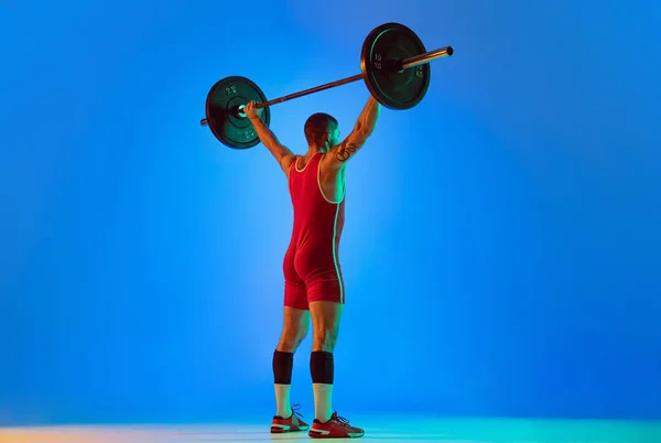 演播室拍摄的年轻男子穿着红色运动服，穿着杠铃隔离蓝色背景的霓虹灯进行运动。体育、举重、力量、成就概念 — 图库照片