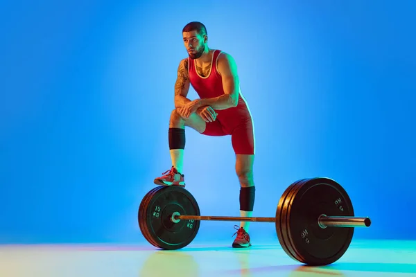 Studio shot van jonge man in rode sportkleding oefenen met lange halter geïsoleerde blauwe achtergrond in neon. Sport, gewichtheffen, kracht, prestaties concept — Stockfoto