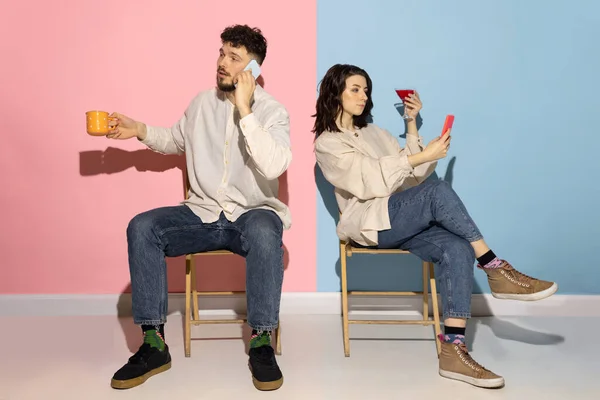 Studio shot van jonge emotionele man en meisje met behulp van telefoons geïsoleerd op blauw en roze trendy kleur achtergrond. Menselijke emoties, zorg, relaties concept — Stockfoto
