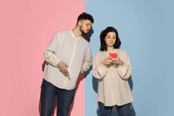 Studio shot van jonge emotionele man en meisje met behulp van telefoons geïsoleerd op blauw en roze trendy kleur achtergrond. Menselijke emoties, zorg, relaties concept — Stockfoto
