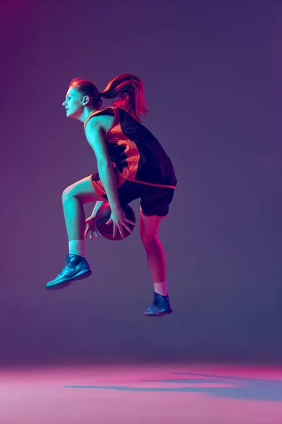 Portret van jong meisje, basketbalspeler dribblig bal in sprong geïsoleerd over verloop roze paarse achtergrond in neon — Stockfoto