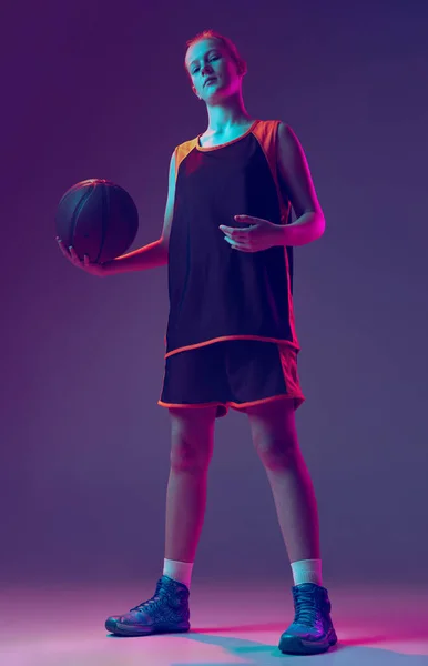 Portret van meisje, tiener, basketbalspeler in uniform poserend met bal op schouder geïsoleerd over hellend roze paarse achtergrond in neon. Kampioen. — Stockfoto