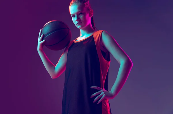 Retrato de niña, adolescente, jugador de baloncesto en uniforme posando con pelota en el hombro aislado sobre el gradiente rosa púrpura fondo en neón — Foto de Stock