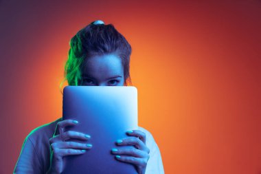 Yüzün yarısını tabletle kaplayan genç bir kızın portresi, neon ışıkta izole edilmiş kırmızı arkaplanda poz veriyor.