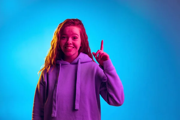 Portrait of young stylish girl rising finger expressing idea isolated over blue studio background in pink neon light — Stock Photo, Image