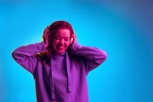 Portret van jong emotioneel meisje vrolijk luisteren naar muziek in hoofdtelefoon geïsoleerd over blauwe studio achtergrond in roze neon licht — Stockfoto
