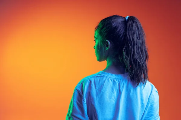 흰색 티셔츠를 입고 네온 지역의 오렌지색 배경 위에서 고립된 포니테일을 하고 있는 어린 소녀의 뒷모습 — 스톡 사진