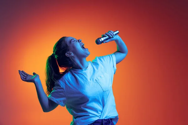 Portrét mladé dívky emocionálně zpívající v mikrofonu, pózující izolované přes gradient červené pozadí v neonovém světle — Stock fotografie