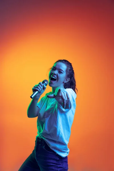 Portret van een jong meisje dat emotioneel zingt in een microfoon, geïsoleerd poserend over een hellende rode achtergrond in neonlicht. Muziek levensstijl — Stockfoto