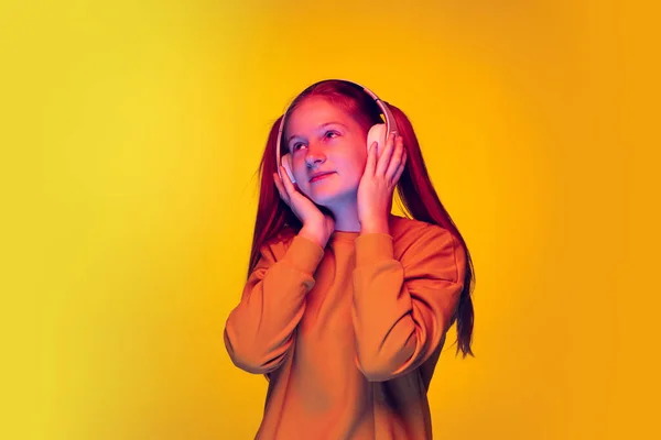 二つのポニーテールを持つ若い女の子の肖像は、ネオンライトで黄色の背景の上に隔離されたヘッドフォンで音楽を聴く — ストック写真