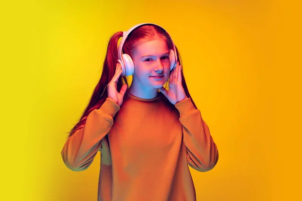 Portret pięknej nastolatki słuchającej muzyki w słuchawkach, pozującej na żółtym tle studia w neonowym świetle — Zdjęcie stockowe