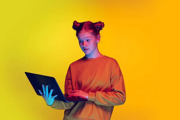 Portrét dospívající dívky dívající se na notebook, pózující izolované žluté pozadí v neonovém světle — Stock fotografie