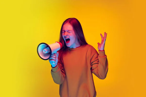 Half-length portrait of young emotive girl expressivelly shouting in megaphone isolated over yellow studio background in neon light