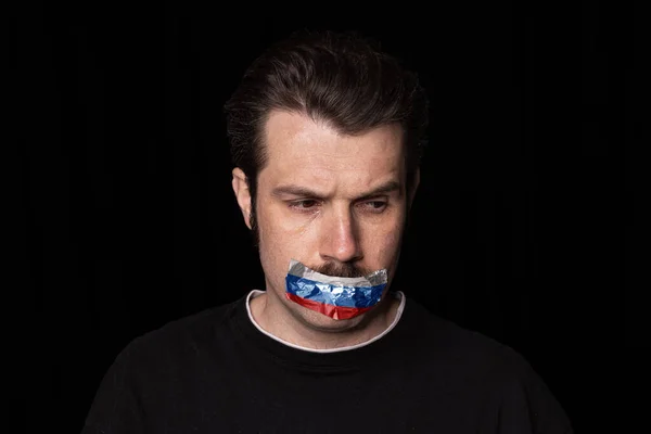 Konzeptporträt eines jungen traurigen Mannes mit dreifarbigem Klebeband über dem Mund, isoliert auf dunklem Hintergrund. Zensur, Konzept der Meinungsfreiheit. — Stockfoto