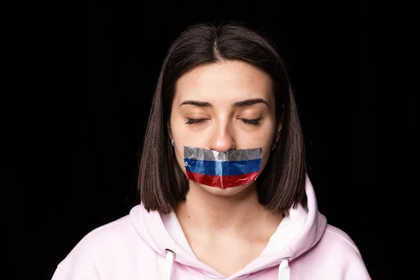Portret młodej zdenerwowanej dziewczyny zamkniętych oczu i trzy kolory taśmy klejącej na ustach odizolowanych na ciemnym tle. Cenzura, koncepcja wolności słowa. — Zdjęcie stockowe