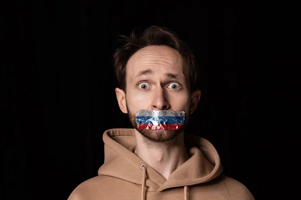 Nahaufnahme Porträt eines jungen emotionalen Mannes mit dreifarbigem Klebeband über dem Mund, isoliert auf dunklem Hintergrund. Zensur, Konzept der Meinungsfreiheit. — Stockfoto