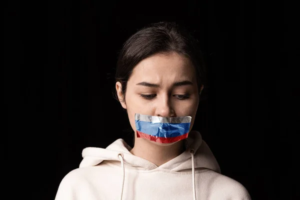 Halbbeiniges Porträt eines jungen emotionalen aufgebrachten Mädchens mit dreifarbigem Klebeband über dem Mund, isoliert auf dunklem Hintergrund. Zensur, Konzept der Meinungsfreiheit. — Stockfoto