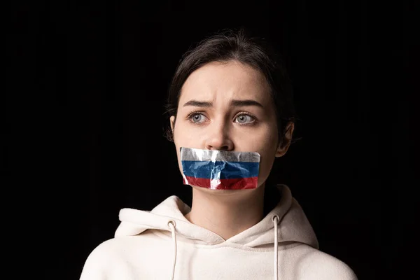 Half-legth portrait of young emotional upset girl with three colors duct tape over her mouth isolated on dark background. Censorship, freedom of speech concept.