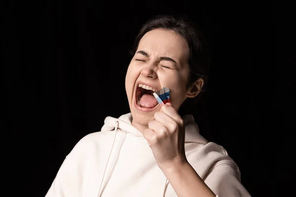 Portrait demi-jambe de jeune fille émotionnelle arrache trois couleurs ruban adhésif conduit de sa bouche isolée sur fond sombre. Censure, concept de liberté d'expression. — Photo