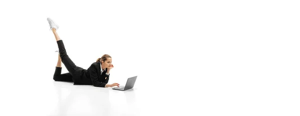 身着正式黑色西服的女商人躺在地板上，用笔记本电脑在白色的工作室背景上打字 — 图库照片