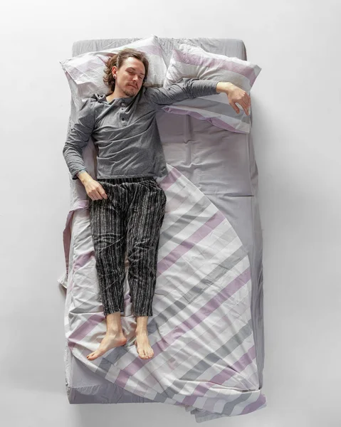 Junger Mann in grauer Hauskleidung, Schlafanzug im großen Bett. Konzept der Gesundheit, Inneneinrichtung, entspannte Zeit, Familie, Träume. Ansicht von oben — Stockfoto