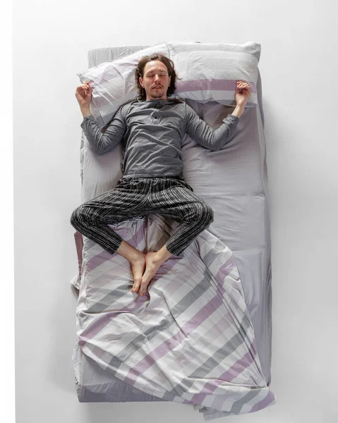 Junger Mann in grauer Hauskleidung, Schlafanzug im großen Bett. Konzept der Gesundheit, Inneneinrichtung, entspannte Zeit, Familie, Träume. Ansicht von oben — Stockfoto