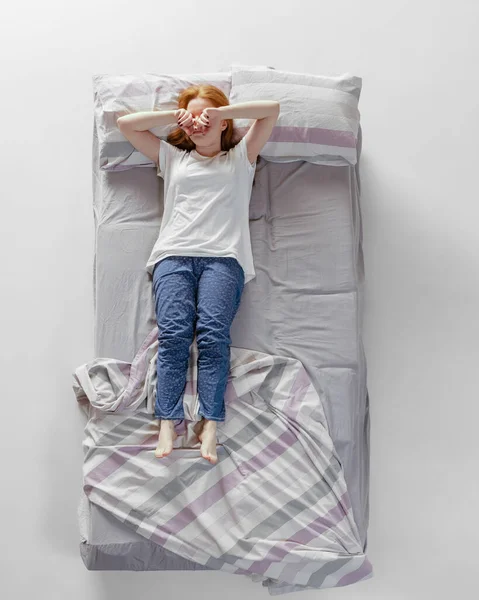 Вид с воздуха на рыжую молодую спящую девушку в хлопчатобумажной одежде, лежащую в огромной серой кровати. Здоровья, домашнего интерьера, отдыха, семьи, мечты — стоковое фото