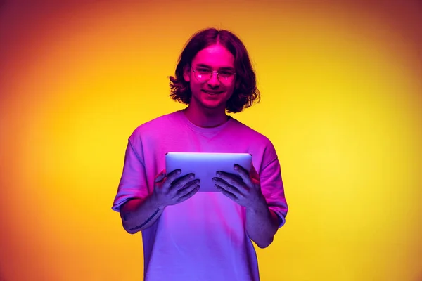 Portrait of young man with long hair using digital gadget isolated on orange background in purple neon light. Concept of emotions, beauty, fashion