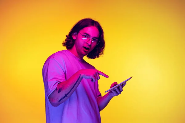 Portrait of young man with long hair using digital gadget isolated on orange background in purple neon light. Concept of emotions, beauty, fashion