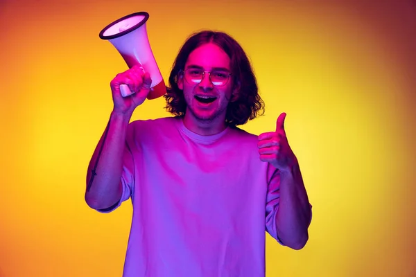 Un giovane bell'uomo in t-shirt bianca che grida al megafono isolato su sfondo arancione in luce al neon. Concetto di emozioni, moda, gioventù — Foto Stock