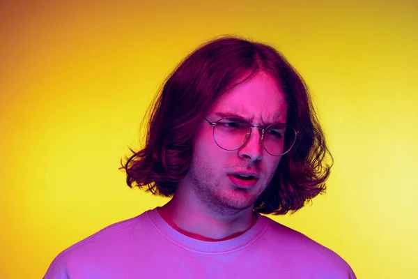 Wygląda na wkurzonego. Portret młodego człowieka, student w luźnych ciuchach odizolowany na pomarańczowym tle w fioletowym neonowym świetle. Pojęcie emocji — Zdjęcie stockowe