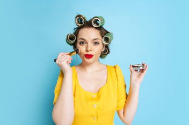 Genç duygusal kızın komik portresi, kafasında bigudiler olan ev hanımı mavi stüdyo arka planında izole edilmiş. Duygular, kadın hakları, güzellik, aile