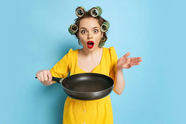 Comic portrait of young emotional girl, housewife with curlers on her head holding pan isolated on blue studio background. Emotions, female rights, beauty, family — Stock Photo, Image