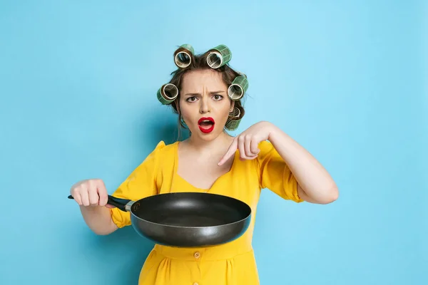 Surprised young girl, housekeeper with curlers on her head holding pan isolated on blue studio background. Emotions, female rights, beauty, family — Stock Photo, Image