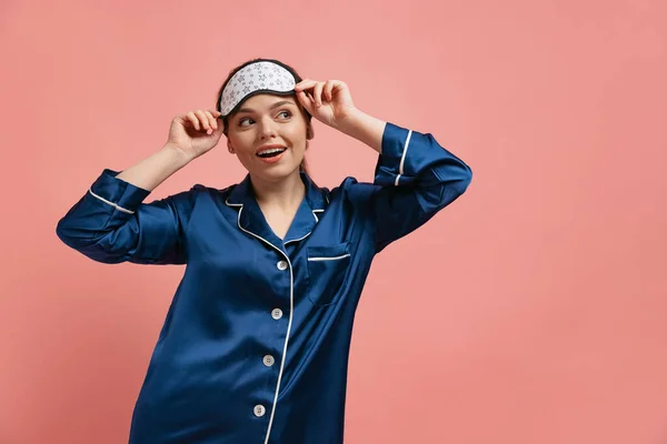 Halblanges Porträt einer jungen, fröhlichen Frau im seidenen Pyjama mit Schlafmaske, die vor rosa Studiohintergrund posiert — Stockfoto