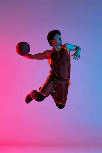 Jovem homem enérgico jogando basquete isolado no fundo do estúdio gradiente rosa azul em luz de néon. Juventude, hobby, movimento, atividade, conceitos de esporte. — Fotografia de Stock