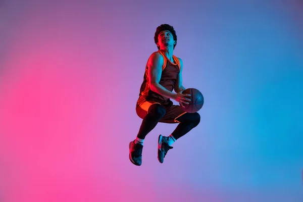 Młody energiczny mężczyzna grający w koszykówkę na gradientowym różowym tle niebieskiego studia w neonowym świetle. Młodzież, hobby, ruch, aktywność, koncepcje sportowe. — Zdjęcie stockowe